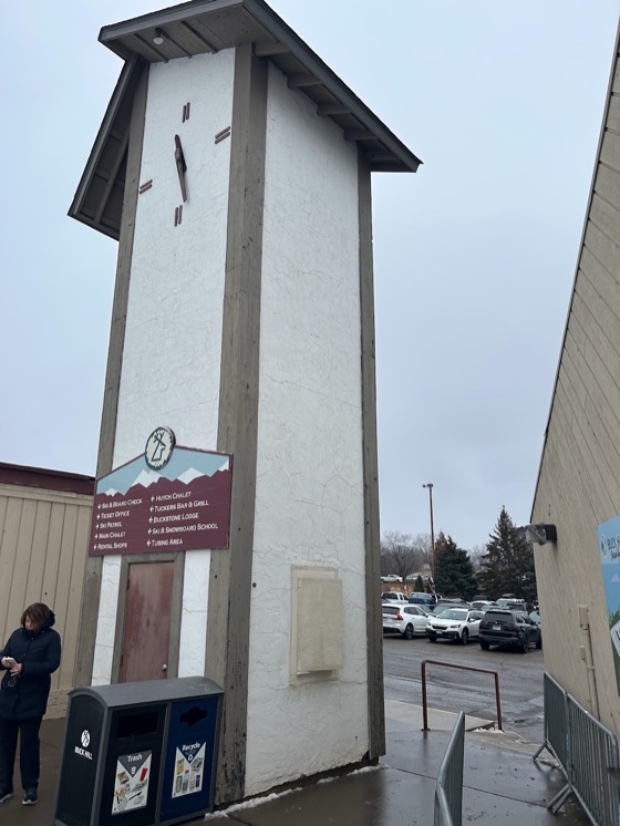 The clock tower at the base of Buck Hill on an overcast day, with a sign listing the ticket office, ski patrol, chalets, the school and the tubing area.