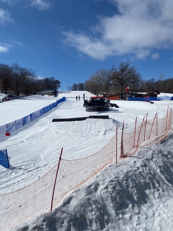 The terrain park at Buck Hill under a blue sky, a groomer parked mid-slope, blue and orange fencing and a black tube feature on the snow.