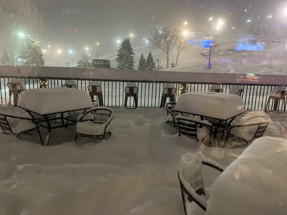 Snowed-in patio tables and chairs at Buck Hill at night, seen through glass, with the lit runs and the chairlift behind.