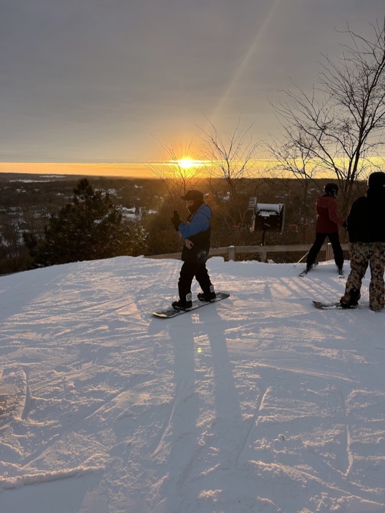 Riders and skiers at the top of Buck Hill at sunset, a snowmaking gun beside them and the sun low over the treeline.