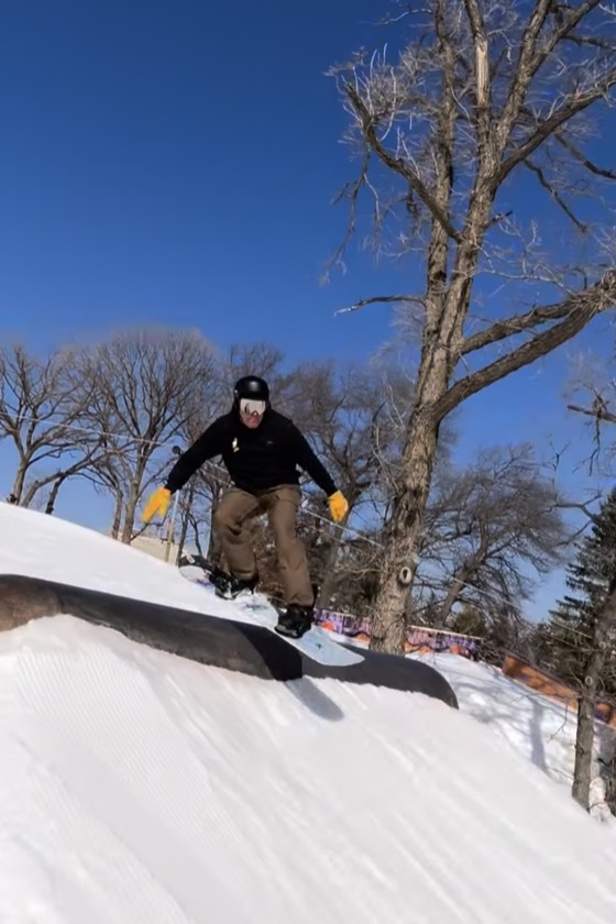 A snowboarder riding a feature in the terrain park at Hyland Hills under a clear blue sky, bare oaks behind.