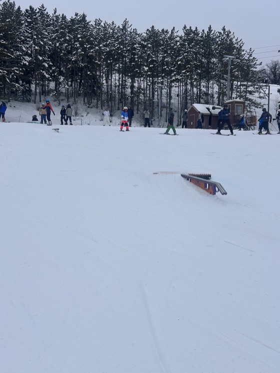 A rail set in the snow at Hyland Hills on a flat grey day, a group waiting at the top by the lift shack and pines behind.