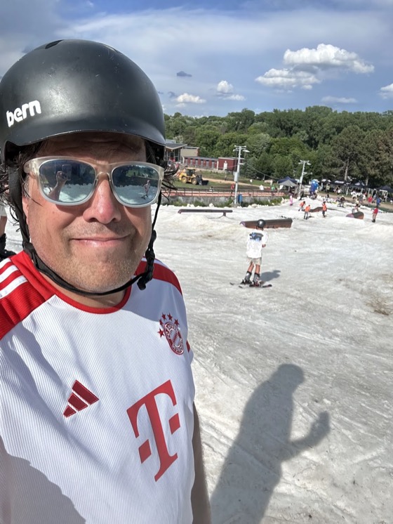 David in a helmet and a football shirt at Hyland Hills in June, riders on cached snow kept over for the event, rails and boxes set across it and green trees behind.