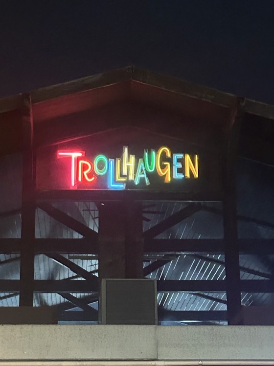A neon Trollhaugen sign in mixed colours on the gable of the wooden chalet at night.
