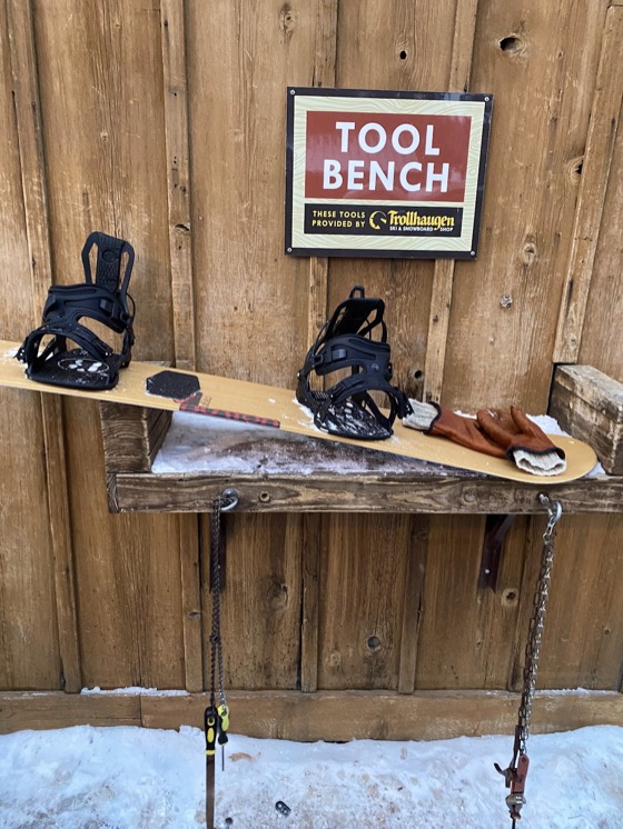 A snowboard laid on a wooden tool bench under a sign reading Tool Bench, tools provided by the Trollhaugen Ski and Snowboard Shop, with leather gloves and a brush beside it.