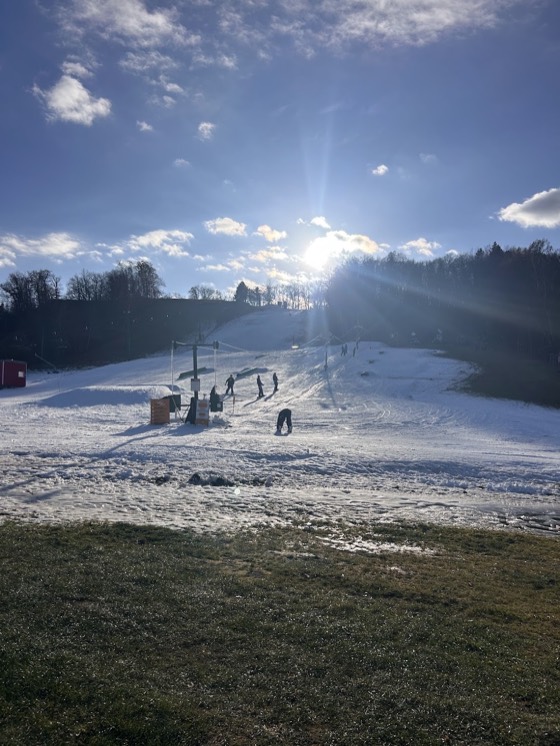 Wild Mountain open on a thin strip of man-made snow at its preseason opening, low sun behind the hill, a few riders on the surface lift and grass showing at the edges.