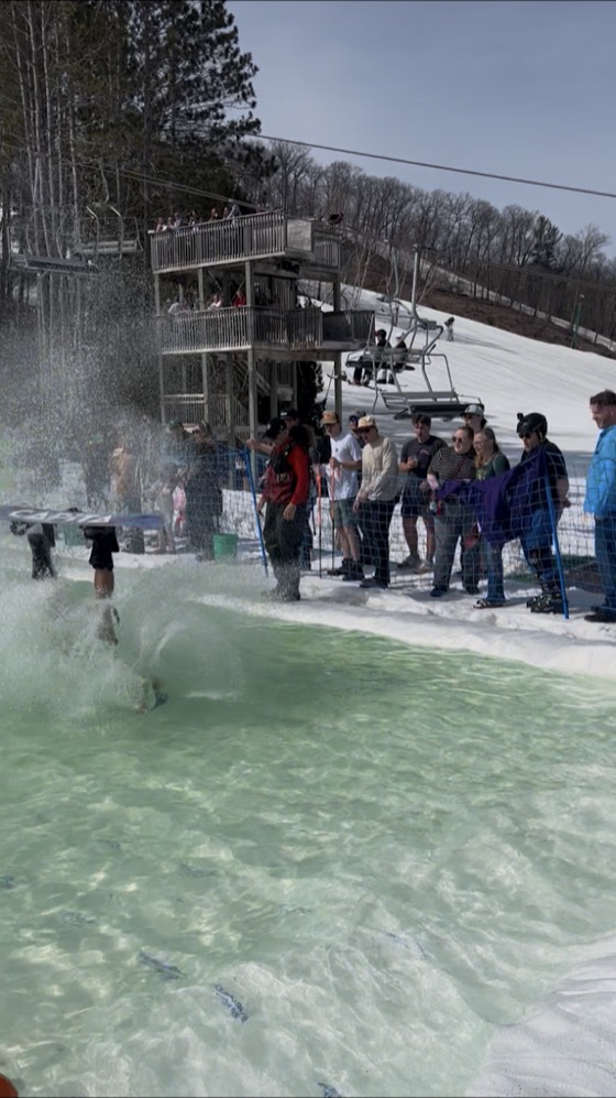 David going down in the pond skim at Wild Mountain, mid-wipeout in a spray of green water, a crowd watching from the fence and the lift tower behind.