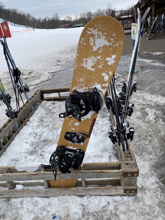 A snowboard standing in a wooden rack outside the chalet at Wild Mountain, skis either side and slush underfoot.